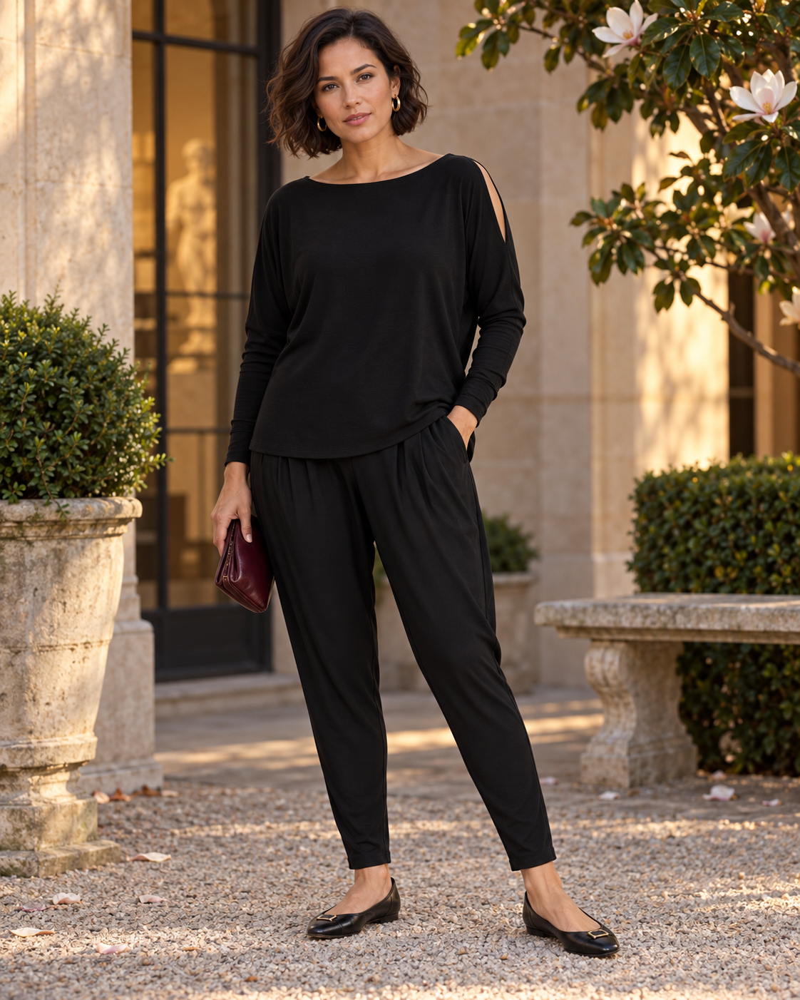 Woman wearing black Paula Ryan Basque pants, an untucked black cold-shoulder top and black ballet flats in a courtyard